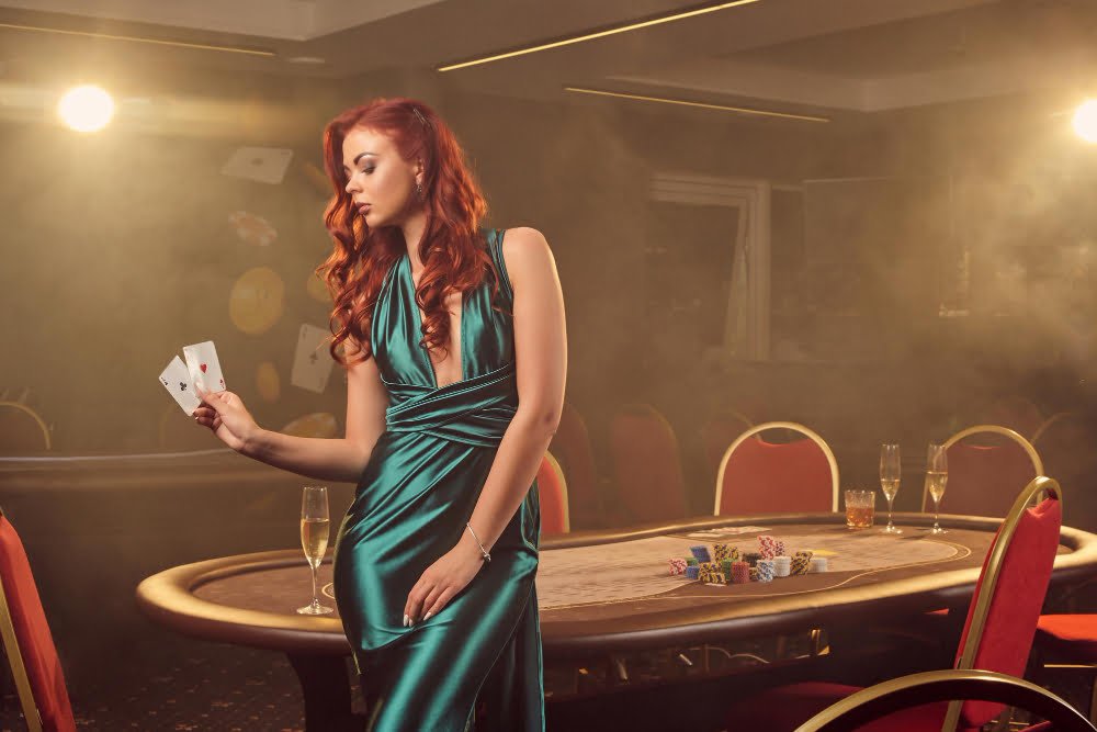 attractive redheaded girl long blue satin dress is posing with two aces her hand against poker table luxury casino passion cards chips alcohol win gambling it is as female enterta
